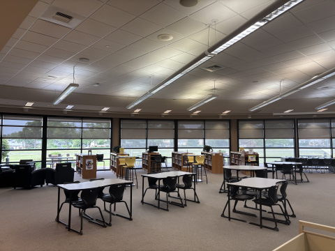 A view of the Oak Forest High School library shows sets of tables and chairs as well as low bookshelves with windows in the distance. 