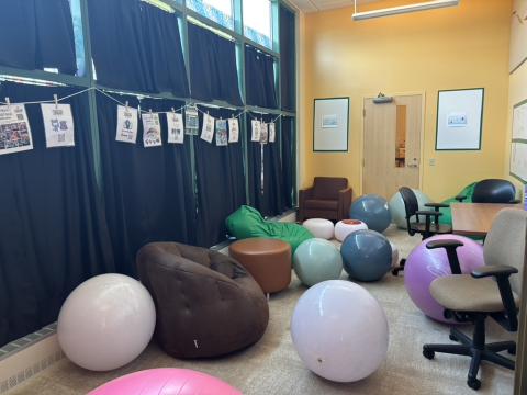 A yellow room with windows covered in blue curtains holds many, varied seating options including office chairs, armchairs, and exercise balls. 