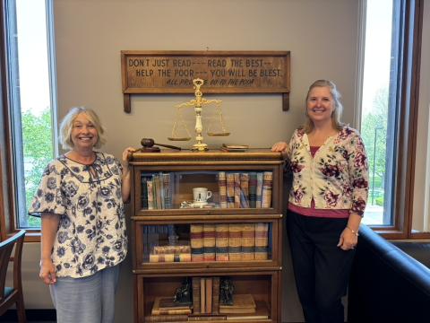 Two librarians stand on either side of a glass front bookshelf holding classic bound, vintage texts. On top of the bookshelf are gold scales and a wooden gavel. A wooden sign on the wall reads, "Don't just read---read the best. Help the poor --- you will be blest. All profits go to the poor."