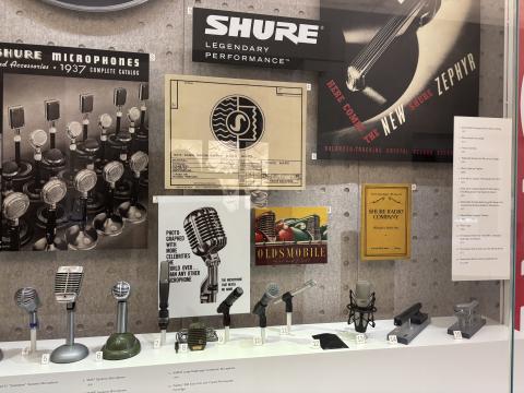 A glass case holds a display of various microphones; numbers corresponding to names printed on the front of the display indicate the model of each. The back of the display features various advertising materials for the Shure Radio Company.