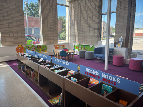 The children's section of the library features board books in display racks and cubbies as well as brightly colored furniture. 