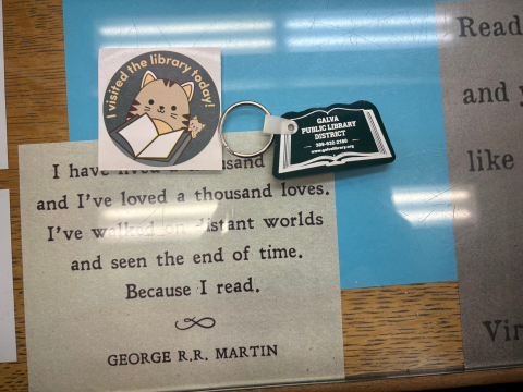 A round sticker featuring a cat and a book reads, "I visited the library today!"  A book-shaped Galva PLD keychain sits to the right of the sticker. All items sit on a desk with a glass cover revealing a piece of parchment which features a George R.R. Martin quote in black text. Portions of the quote are obscured by the sticker. 