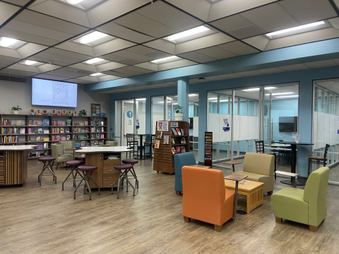 A school library featuring seating areas with mixed types of colorful chairs and stools. In the distance you can see bookshelves and study rooms. 