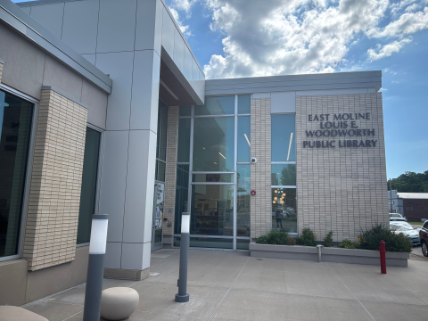 The main entrance to the East Moline Louis E. Woodworth Public Library, a light brick and stone tile building with a concrete walk leading to the front. 