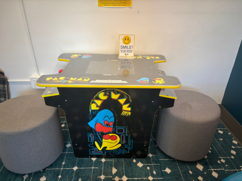 A black, Pac-Man themed table with two grey, padded stools on two sides sits before a white wall. 