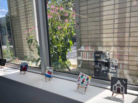 Small-scale paintings sit in tabletop easels on a window ledge overlooking a concrete wall. 
