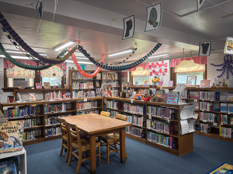 The library's children's section features shelves full of books with displays on top, a table and chairs, as well as colorful decorations hanging from the ceiling and on the walls. 