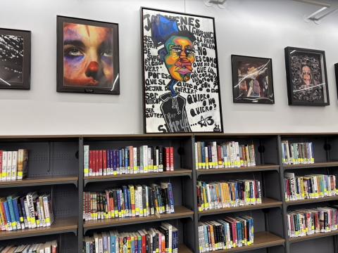 Dark metal bookshelves with walnut shelves hold library books along a white wall. Above the shelves, various colorful artworks are hung in black frames. 