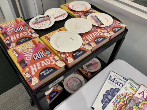 A black media cart holds bedazzled copies of the book Roof Over Our Heads by Nicole Kronzer. Resting on the books are white paper plates holding craft supplies for bedazzling. 