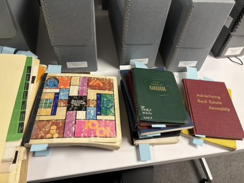 A white table top holds various real estate journals, handbooks, and texts. A stack of ivory file folders is at the left. 