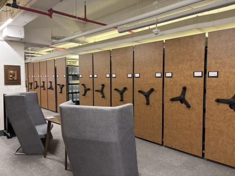 Rows of movable library stacks, each with a wooden end panel and black crank handle, hold grey periodical boxes in the library's collection. 