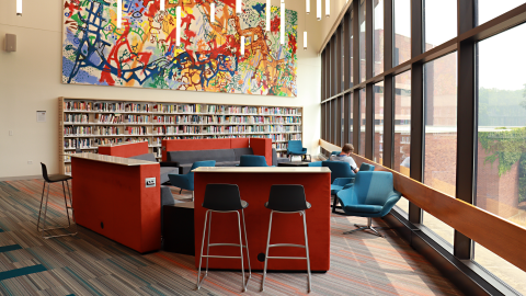 A large abstract painting displayed above a wall of books. Read and blue seating surrounding it. Long lights hang from the ceiling. 